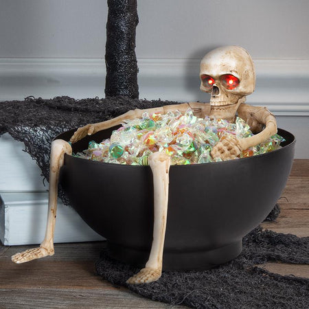 Skeleton figure sitting inside a bowl filled with colorful candy on a Halloween-themed setting.