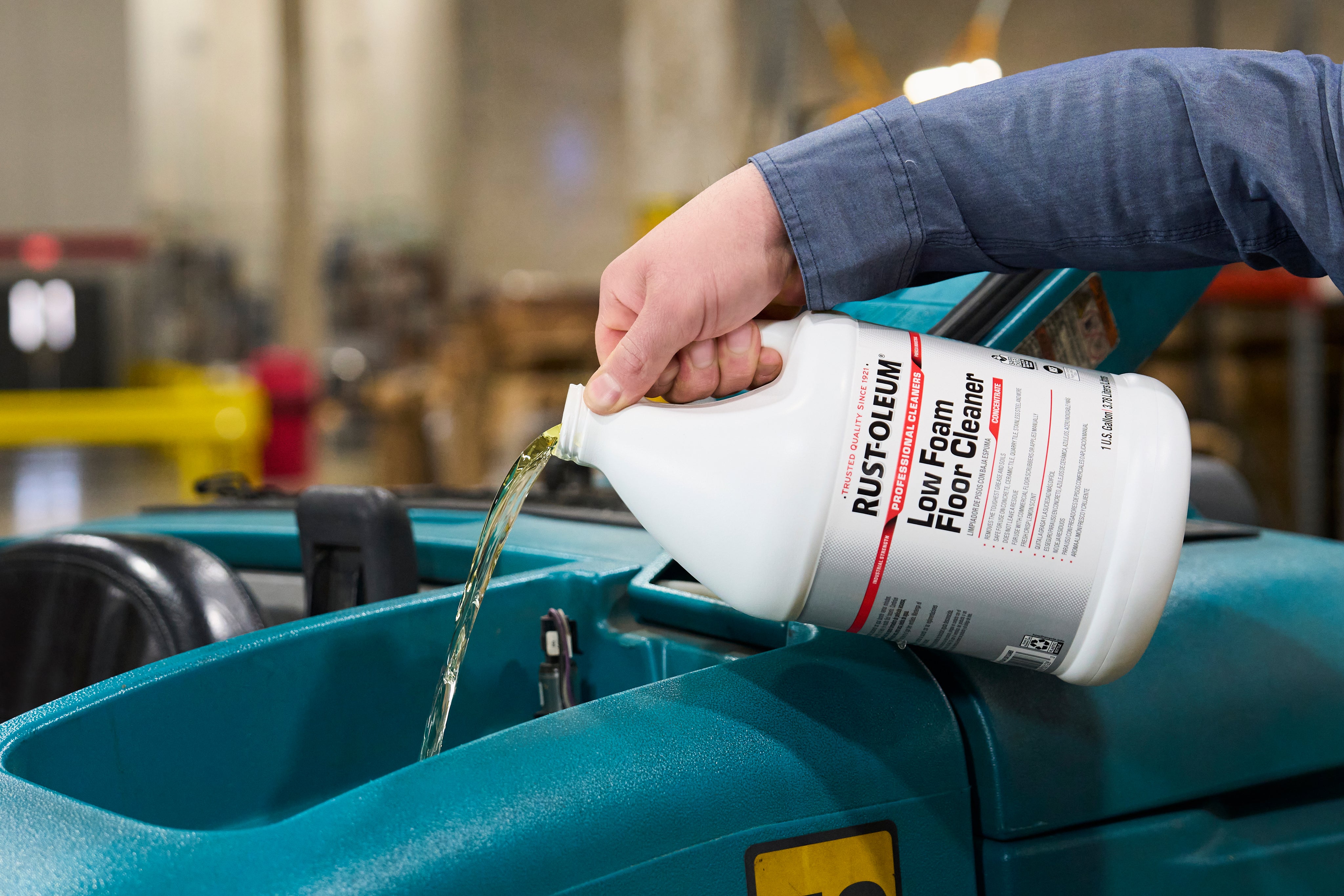 A person in a blue work shirt pours Rust-Oleum Low Foam Floor Cleaner into a teal machine compartment in a workshop setting.