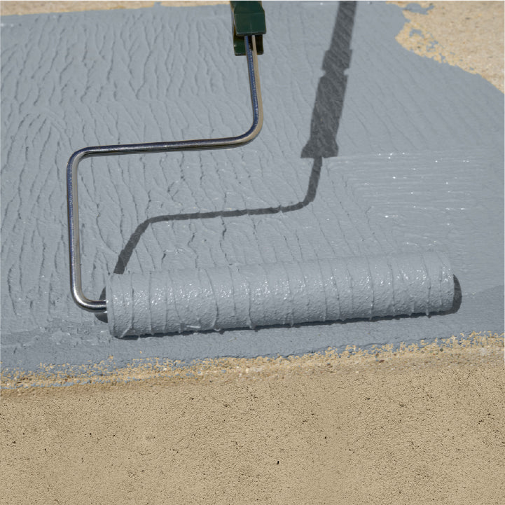 A paint roller applying a thick, textured coat of light gray Safetex Easy Tread paint onto a sandy surface outdoors.