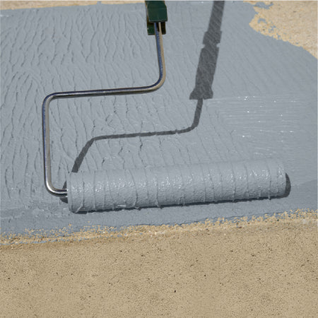 A paint roller applying a thick, textured coat of light gray Safetex Easy Tread paint onto a sandy surface outdoors.