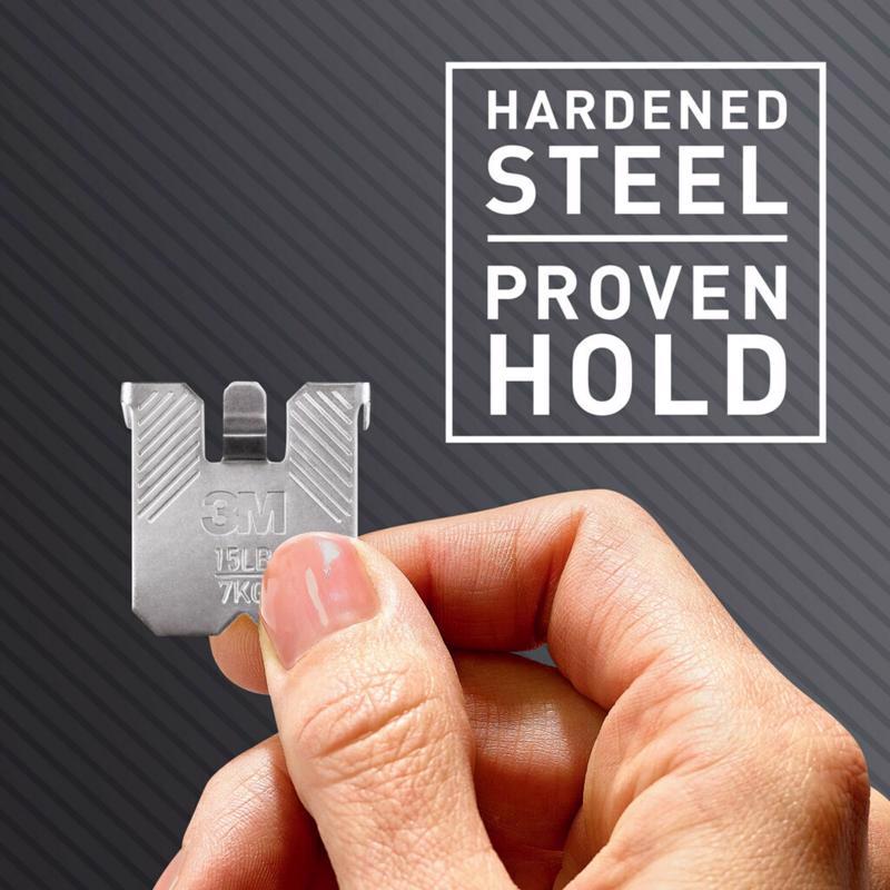 Hand holding a 3M CLAW drywall picture hanger with 'Hardened Steel Proven Hold' text on a dark background