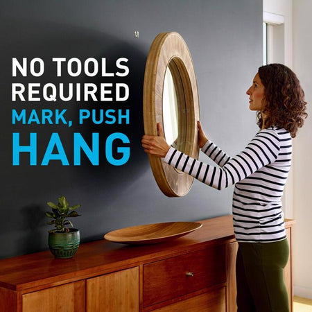 Woman installing a wooden mirror on a wall with text 'No tools required, mark, push, hang'.