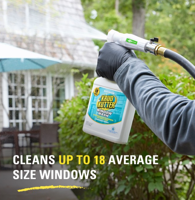 A person wearing a black glove holds a Krud Kutter Window Wash spray bottle while spraying water outdoors. The background shows a blurred house and greenery, with text at the bottom stating 'CLEANS UP TO 18 AVERAGE SIZE WINDOWS'.