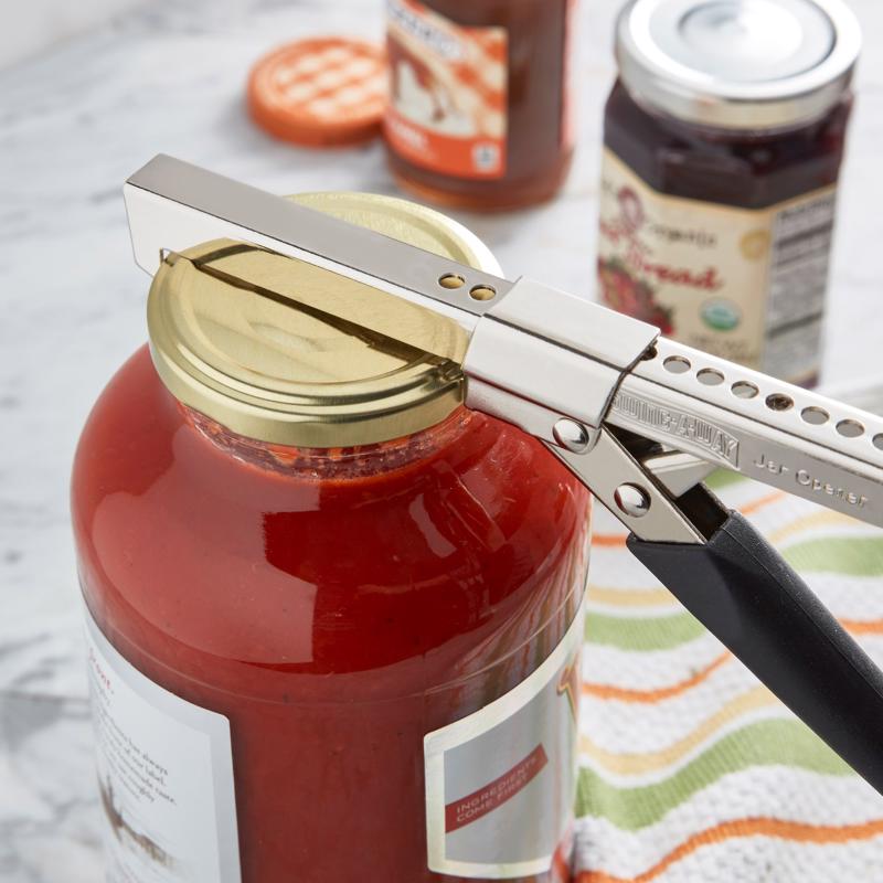 Can opener being used on a red jar with a blurred background of food items.