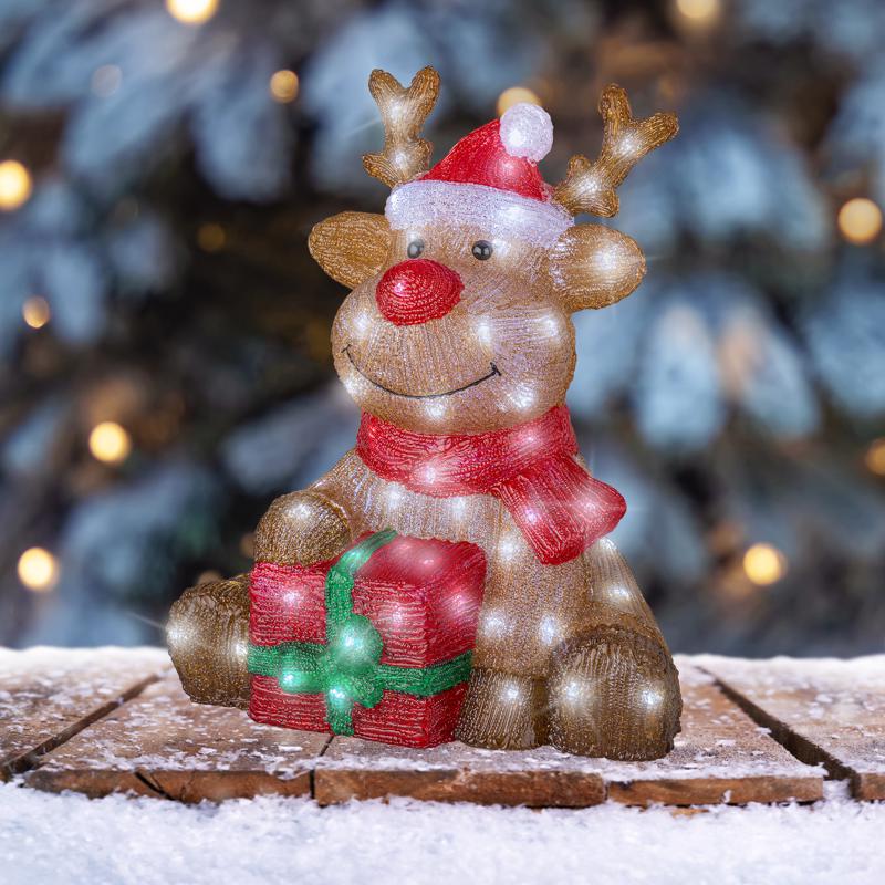 Decorative reindeer figurine with a red nose and gift, set against a blurred Christmas tree background.