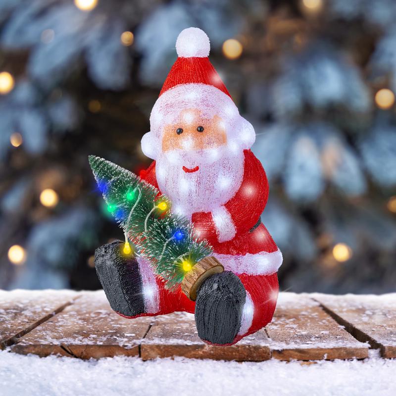 Decorative Santa Claus figurine with a small tree on a snowy surface with blurred Christmas tree and lights in the background.