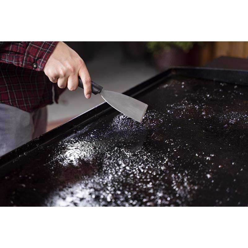 Man using the scraper from the Blackstone 8-Pc Culinary Grill Cleaning Kit 5463 on an outdoor grill