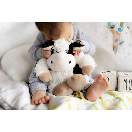 Child holding a Warmies CP-COW-3 Black & White Stuffed Cow toy on a white blanket