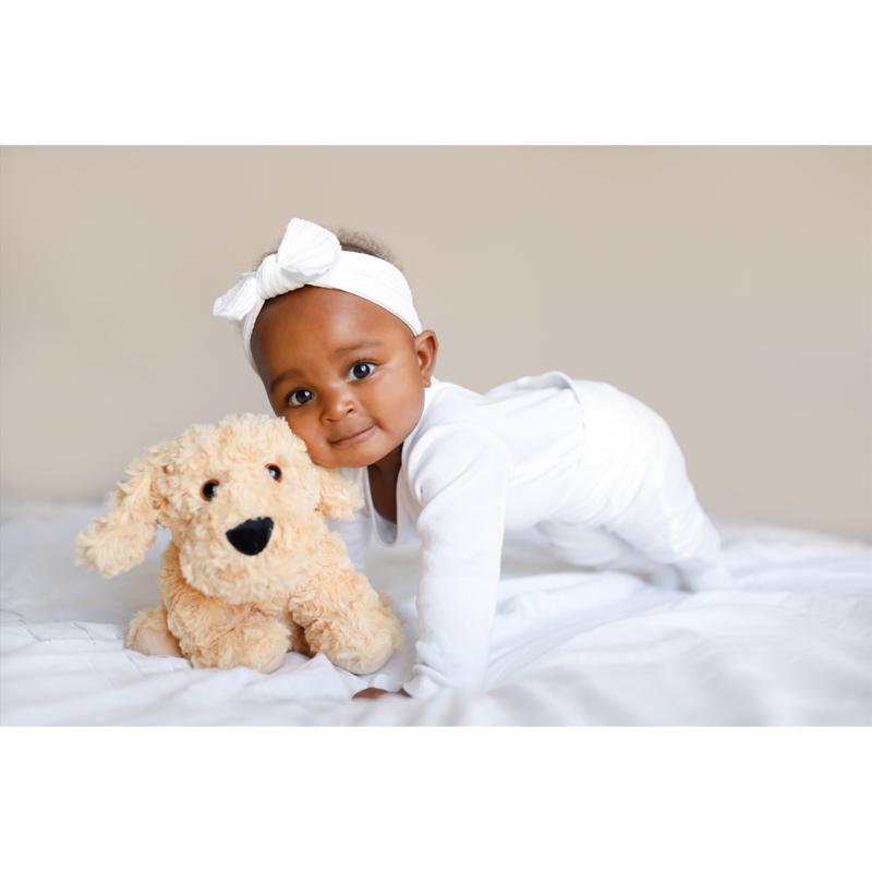 Baby in white outfit with a bow, holding a Warmies CP-DOG-3 Plush Golden Dog toy on a white blanket.