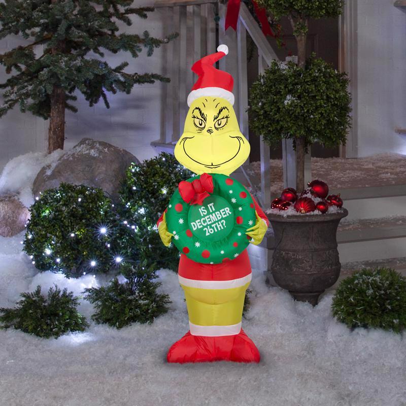 Inflatable Grinch holding a wreath with Christmas text in a snowy outdoor setting.