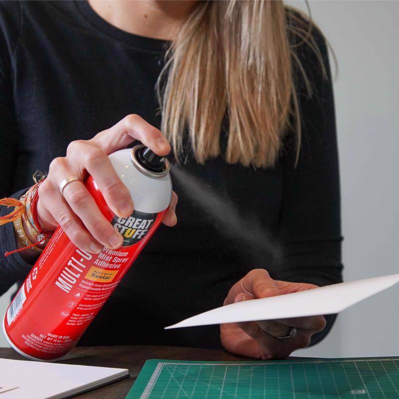 Great Stuff Multi-Use Premium Mist Spray Adhesive being applied to the back of a piece of paper board.
