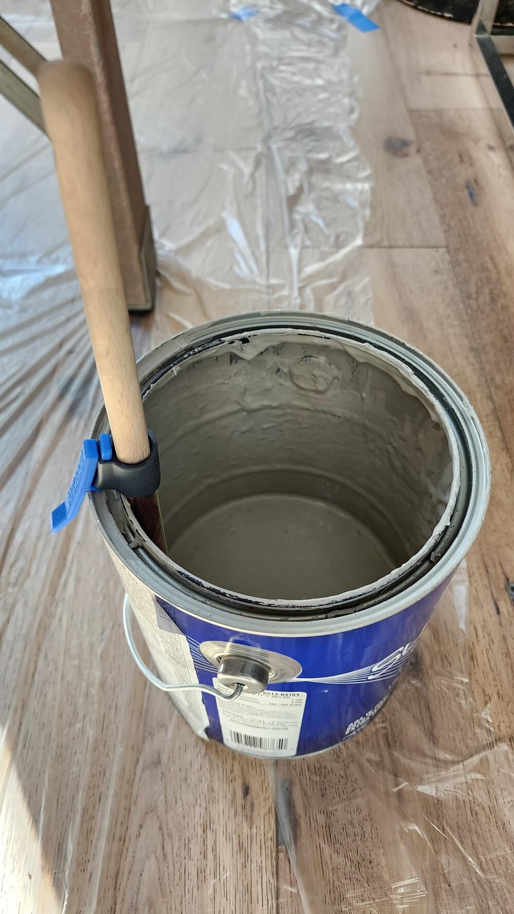 An open blue paint can filled with gray paint, with a paintbrush resting inside and a blue painter's tape loop holding the brush handle. The can sits on a wood floor covered with plastic sheeting.