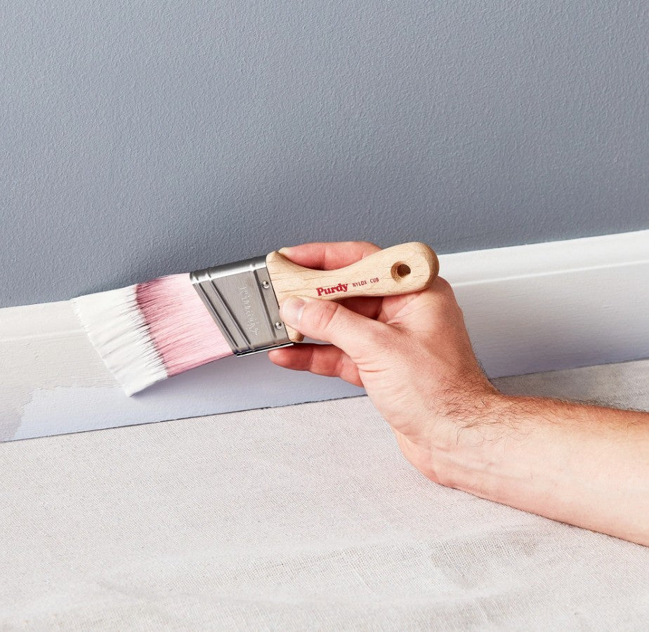 Man holding a Purdy Nylox Cub paint brush applying white paint to a gray wall.