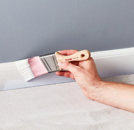 Man holding a Purdy Nylox Cub paint brush applying white paint to a gray wall.
