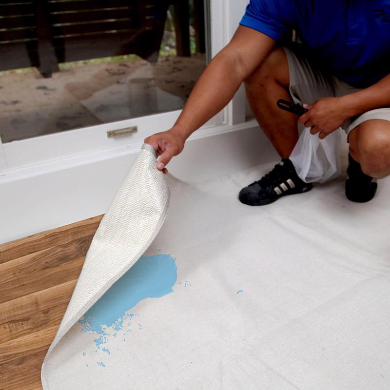 Trimaco Stay Put Canvas Drop Cloth being lifted off a hardwood floor with spilled paint