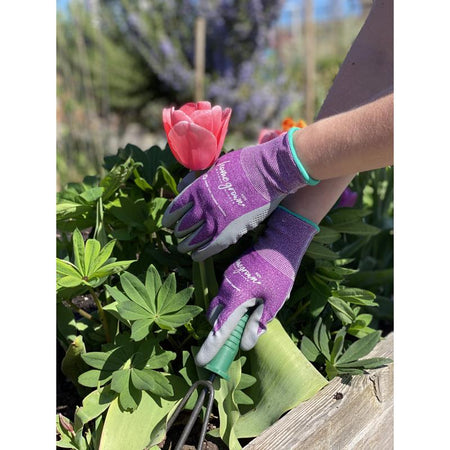 Person wearing gardening gloves and using a tool on plants with a pink flower in the background.